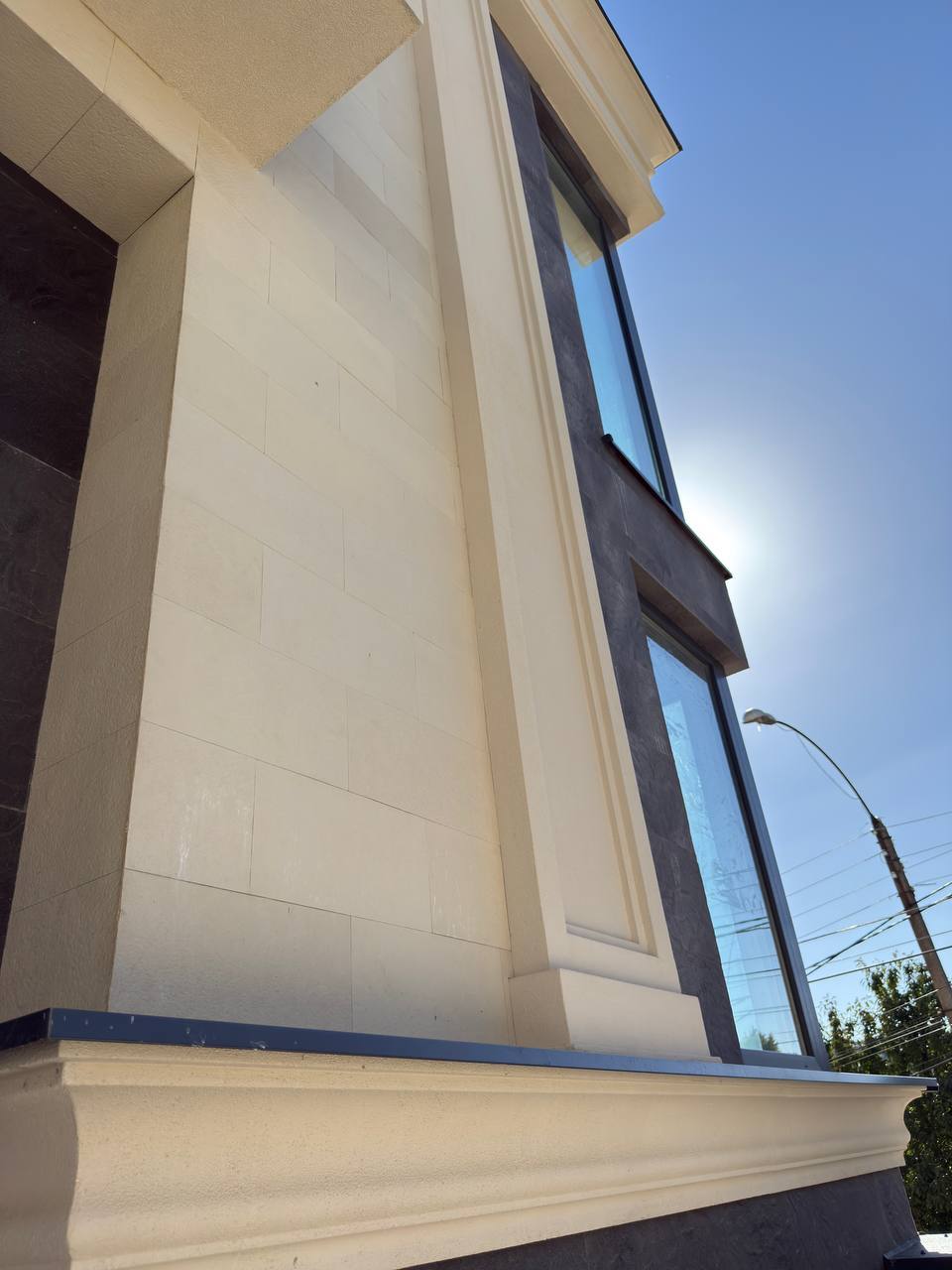Vertical anthracite band and beige pilaster beside a window, bracketed cornice below, shot against the sun