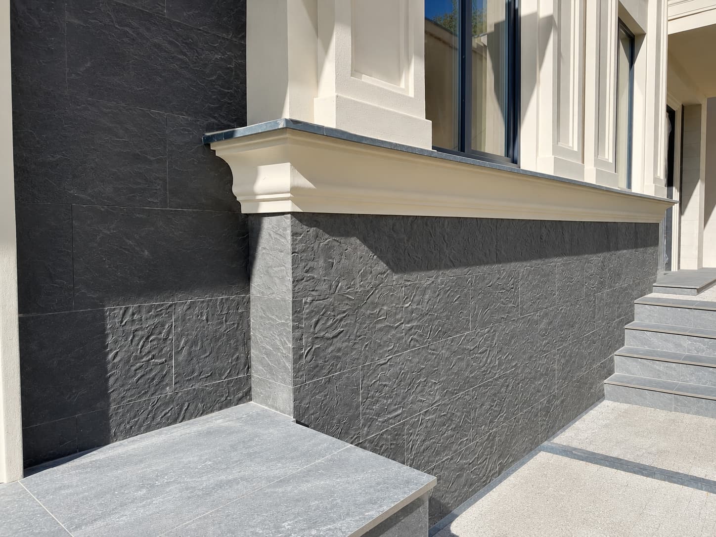 Junction of the bush-hammered beige field and the anthracite split-face plinth below a cornice