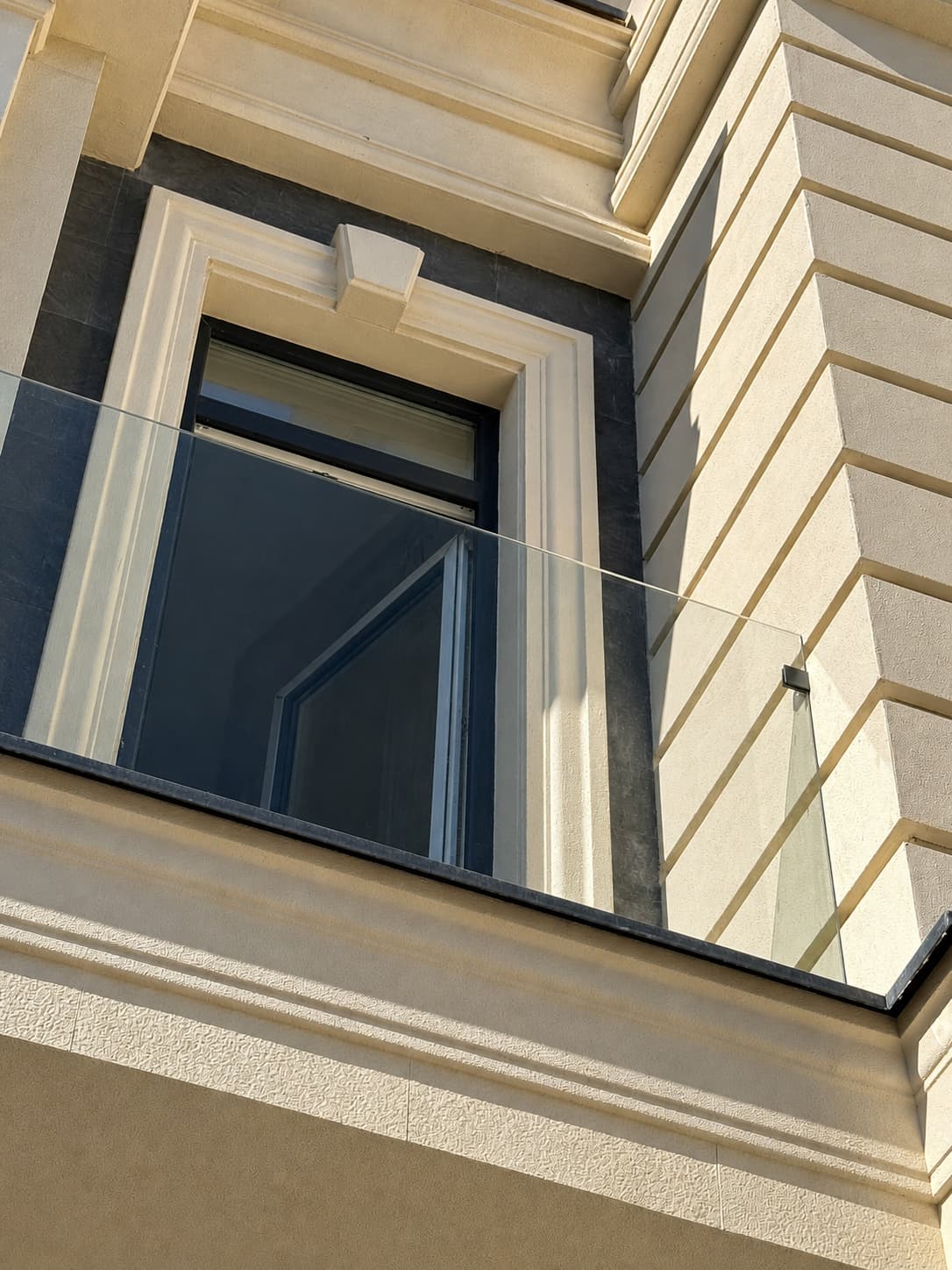 Window with a glass balustrade framed in beige flexible stone above a bracketed cornice